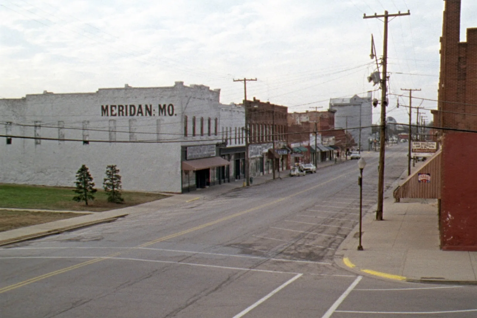 A wide, empty two-block main street of low brick storefronts with awnings and diagonal parking in a small Missouri farm town, with a grain elevator at the far end of the street under an overcast sky.