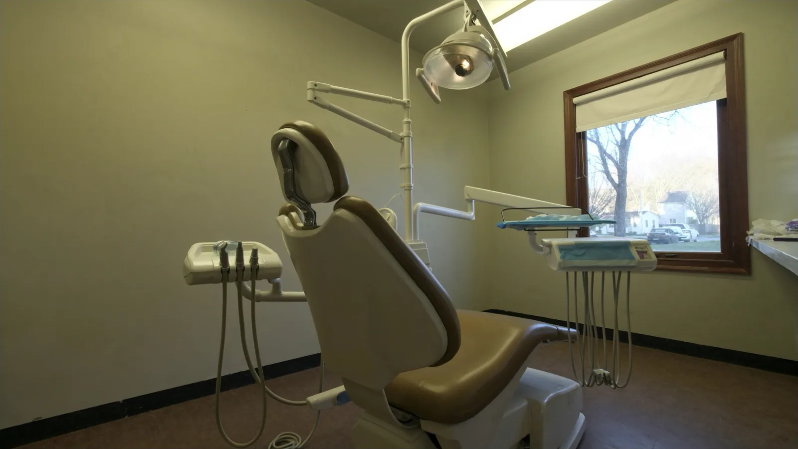 A single dental operatory with a tan vinyl chair, a folded overhead light arm and an instrument tray, beside a window looking onto a residential street with a bare tree, parked cars and a house across the way.