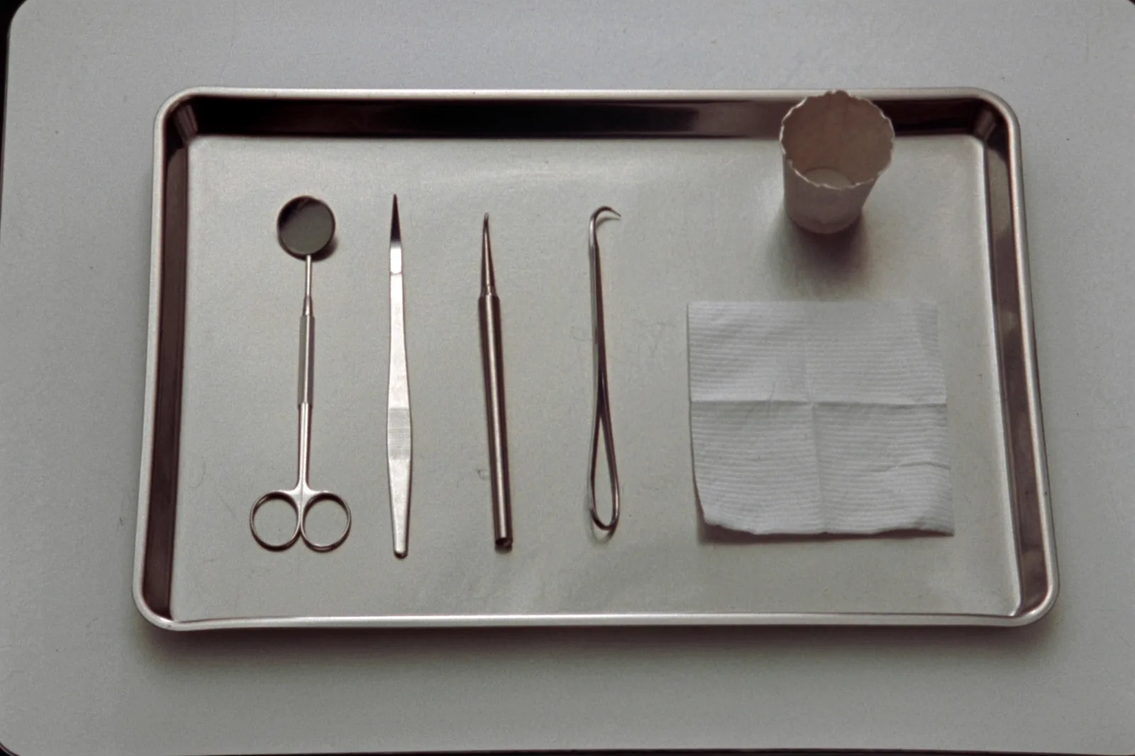 A stainless steel tray photographed from above holding a mouth mirror, tweezers, a probe and a hooked explorer laid out in a row, with a folded gauze square and a small paper cup beside them.