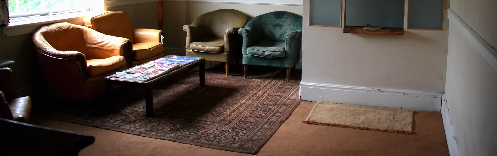Four mismatched armchairs in tan leather, olive and teal velvet around a low table of magazines, on a worn patterned rug beside the base of a reception hatch.